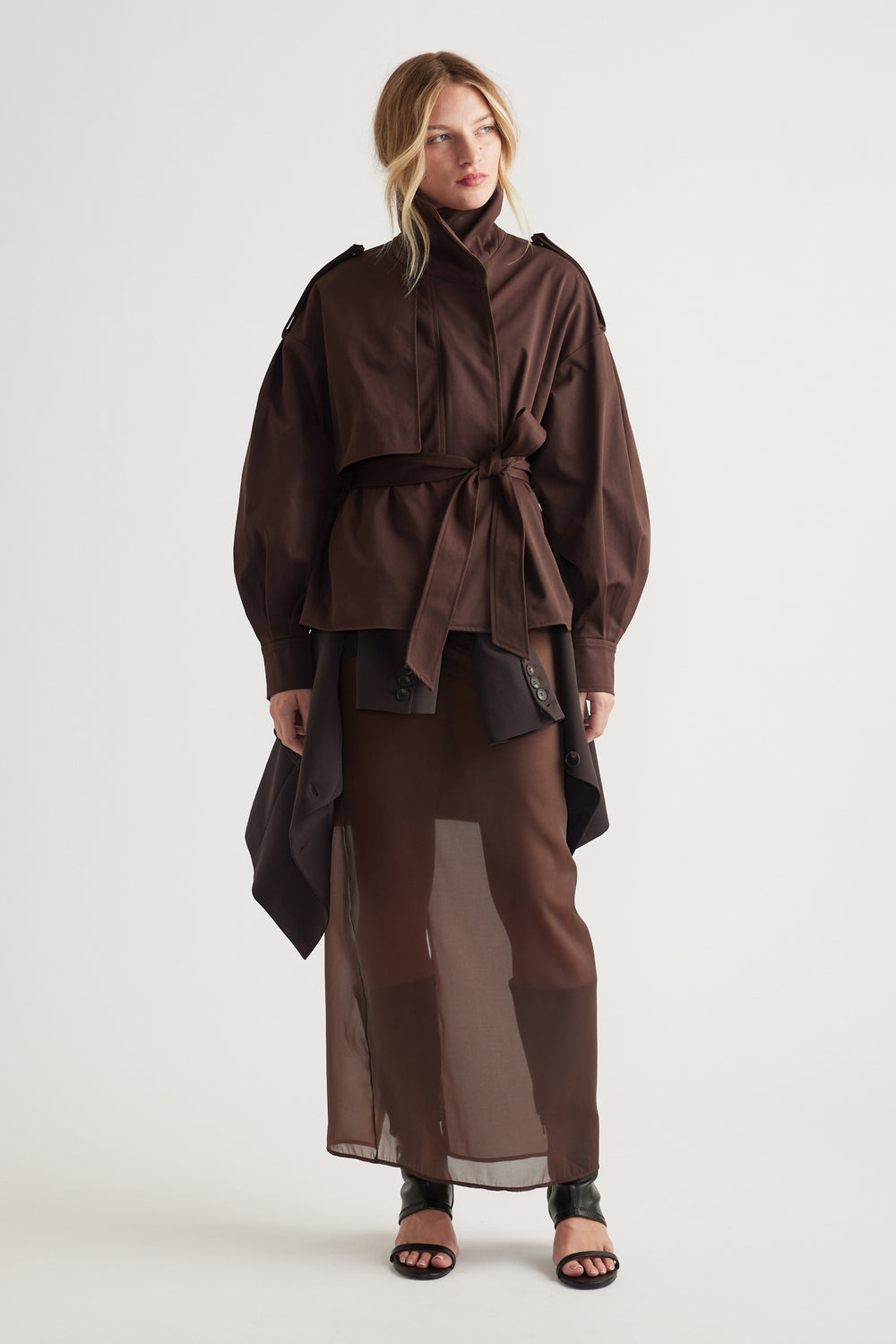 Woman wearing a chocolate brown cropped trench with a waist tie, paired with a sheer skirt and black open-toe heels