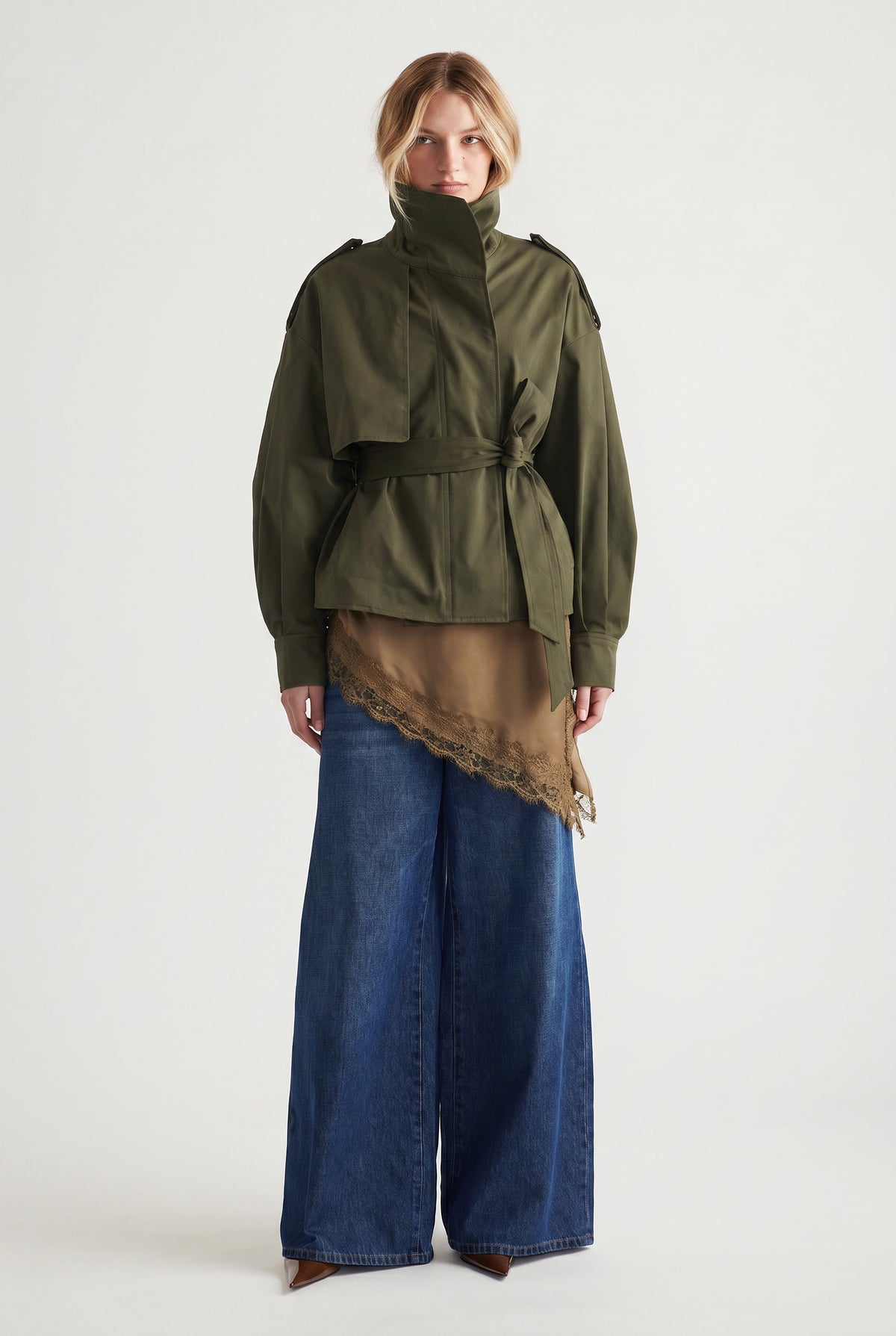 Woman wearing an olive cropped trench with a tie waist, layered over a brown lace-trimmed top and wide-leg denim pants