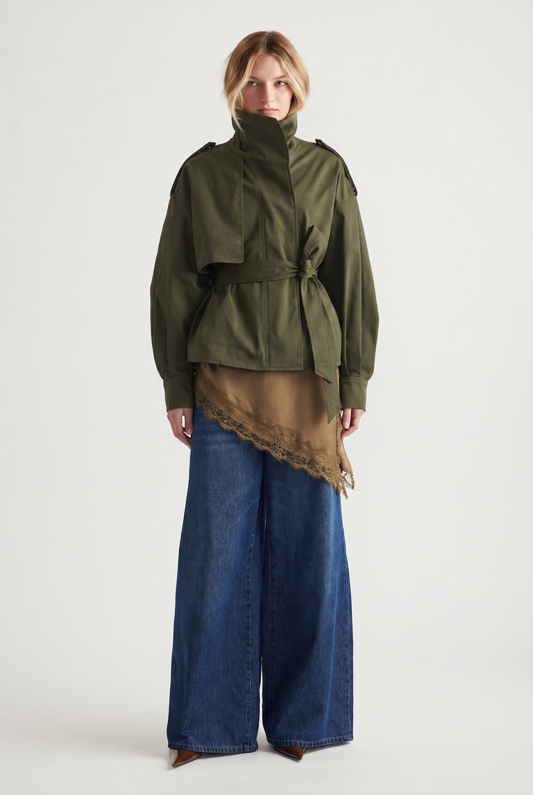 Woman wearing an olive cropped trench with a tie waist, layered over a brown lace-trimmed top and wide-leg denim pants