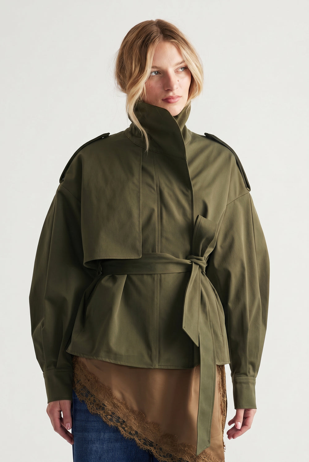 Woman wearing an olive cropped trench with structured shoulders and a tied waist belt, styled casually with a layered skirt.
