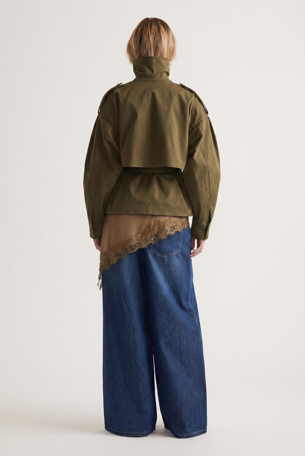 Back view of woman wearing cropped olive green trench jacket over lace-trimmed top and wide-leg denim jeans