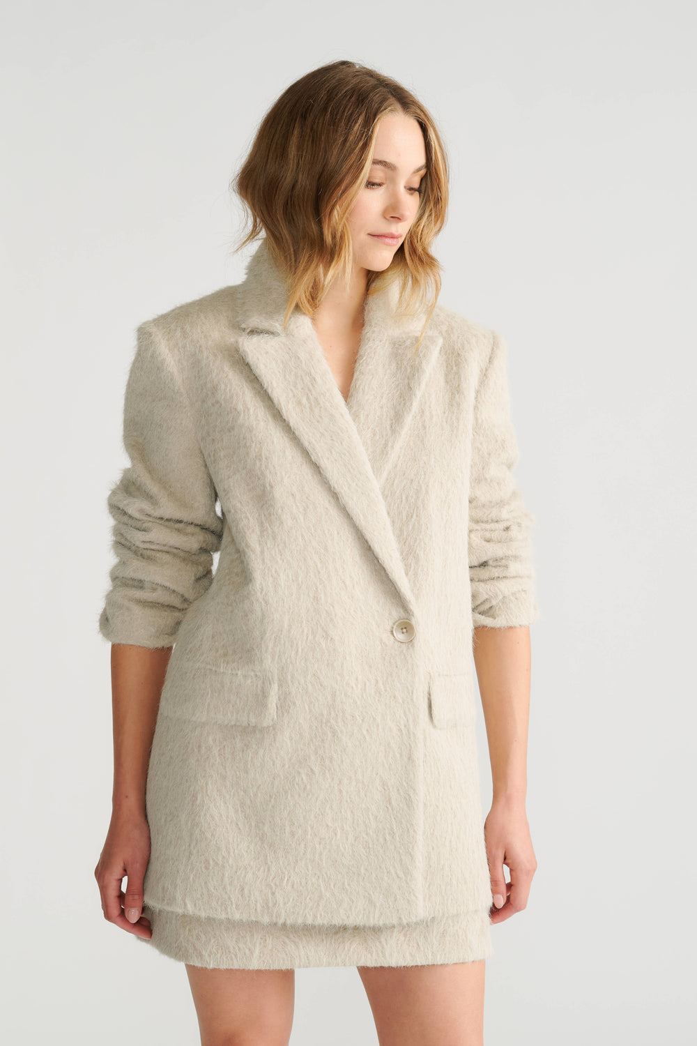 Woman wearing a soft textured oversized beige blazer with ruched sleeves and front flap pockets for a relaxed fit.