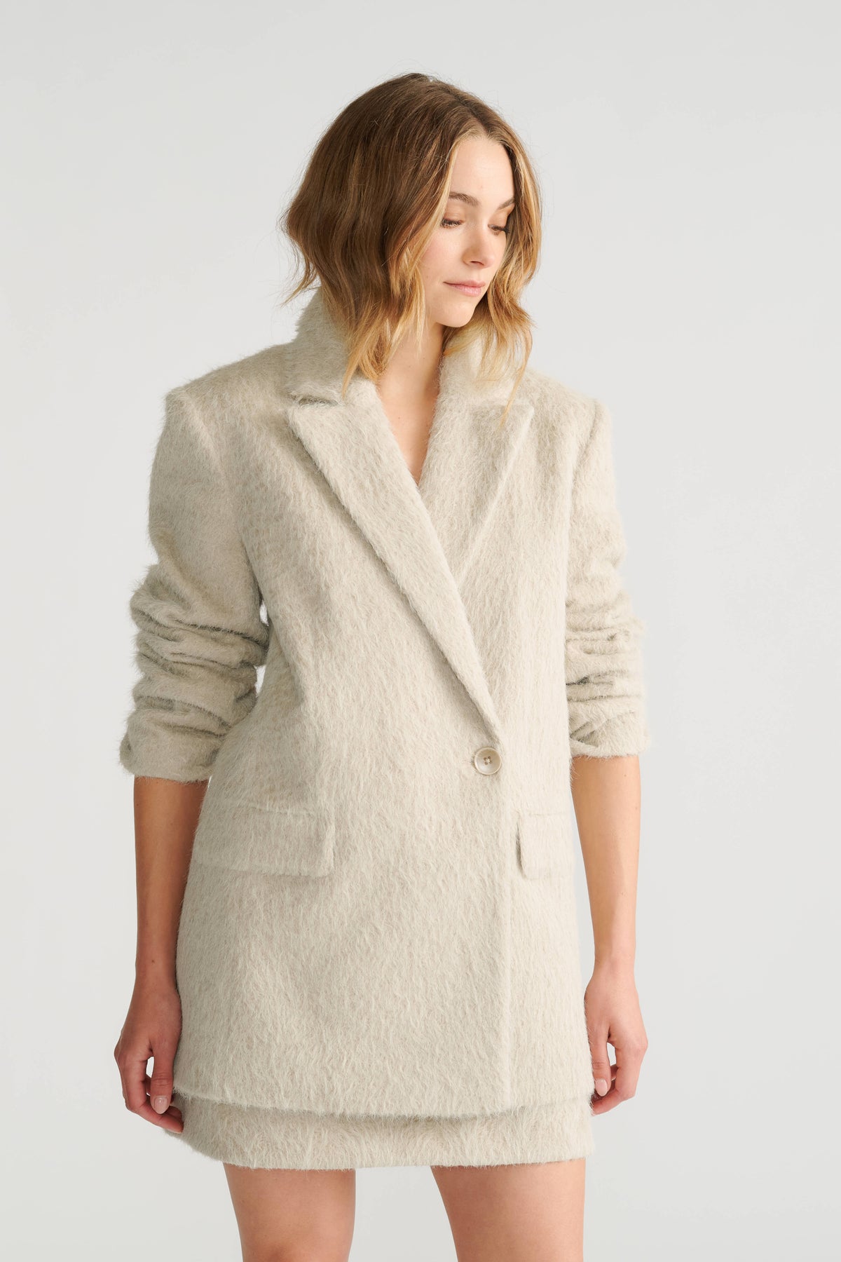 Woman wearing a soft textured oversized beige blazer with ruched sleeves and front flap pockets for a relaxed fit.