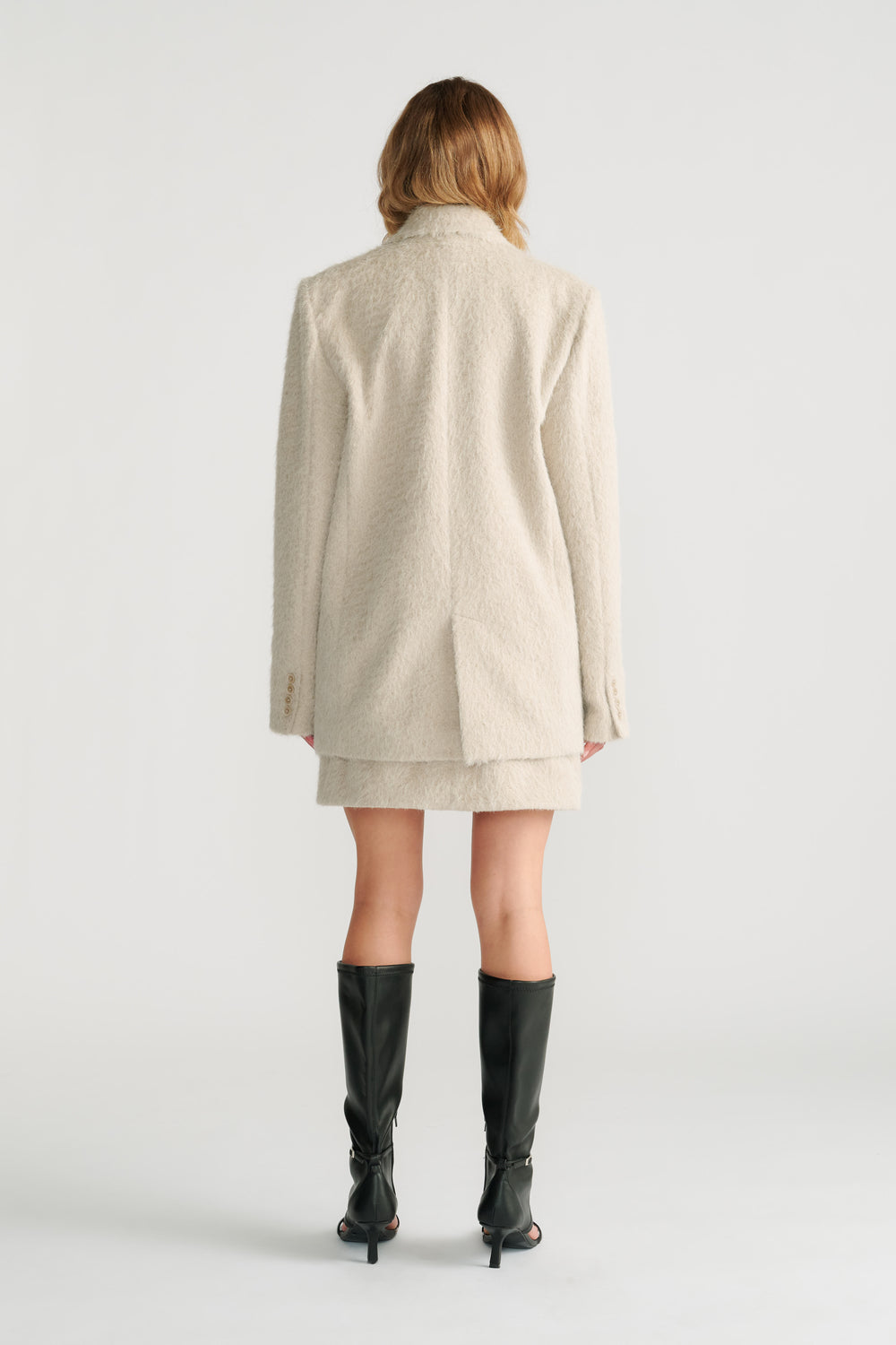 Back view of a woman wearing a textured light stone oversized blazer with matching skirt and black heeled boots.