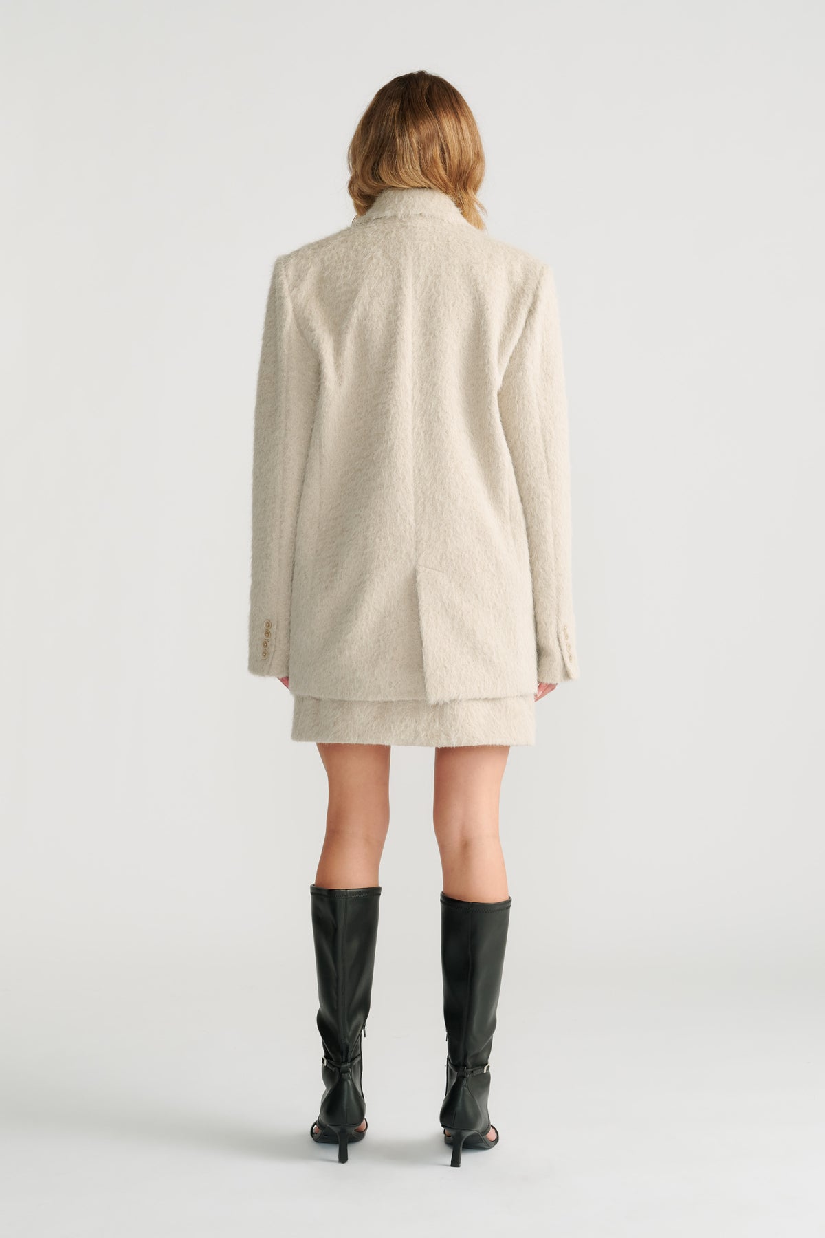 Back view of a woman wearing a textured light stone oversized blazer with matching skirt and black heeled boots.