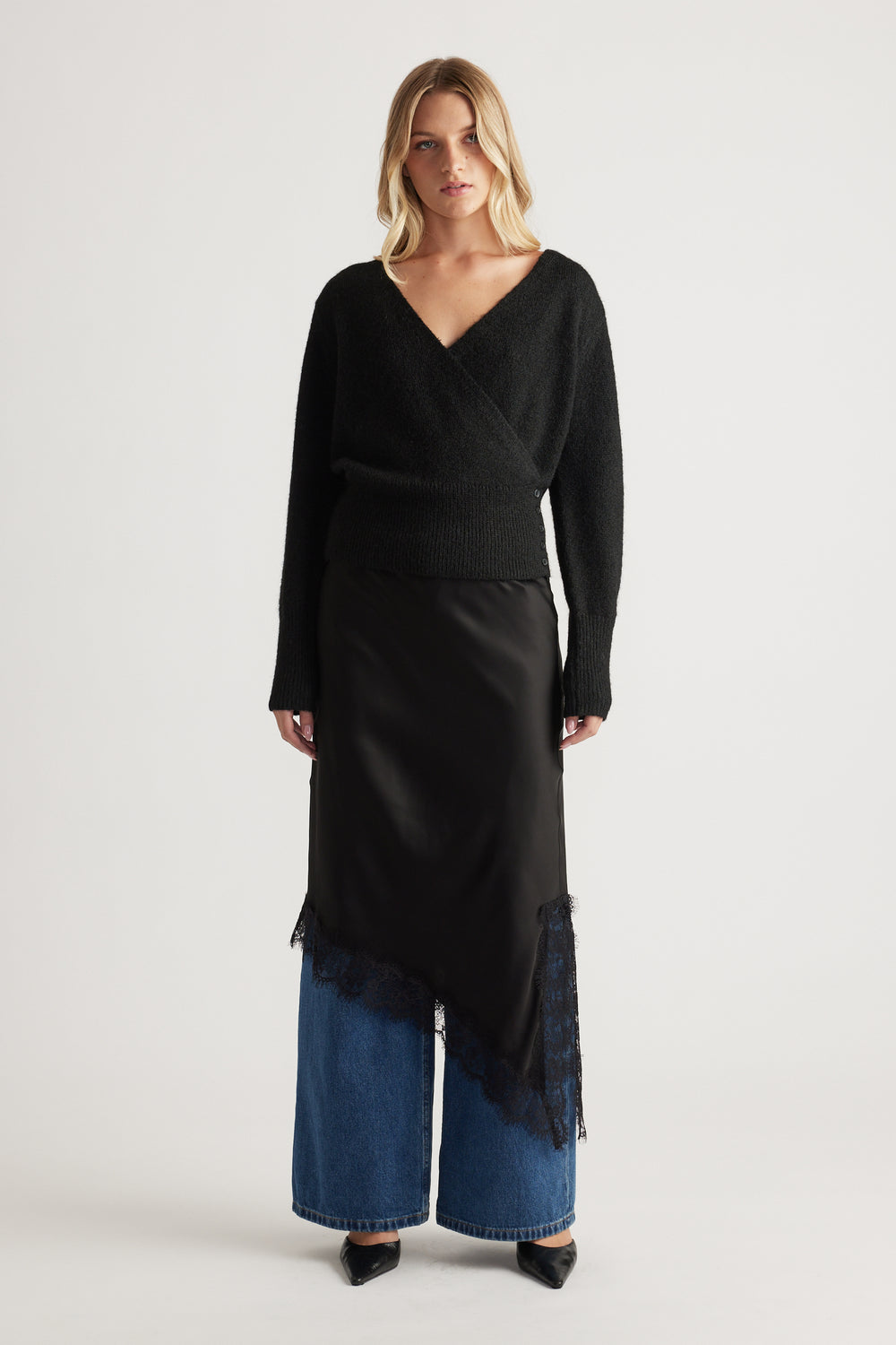 Woman wearing a cozy black crossover knit paired with a silky asymmetrical skirt layered over wide-leg denim jeans.