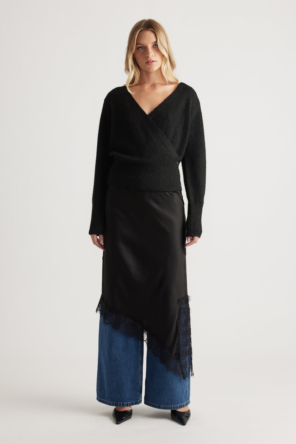 Woman wearing a cozy black crossover knit paired with a silky asymmetrical skirt layered over wide-leg denim jeans.