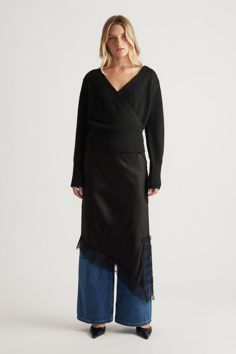 Woman wearing a cozy black crossover knit paired with a silky asymmetrical skirt layered over wide-leg denim jeans.