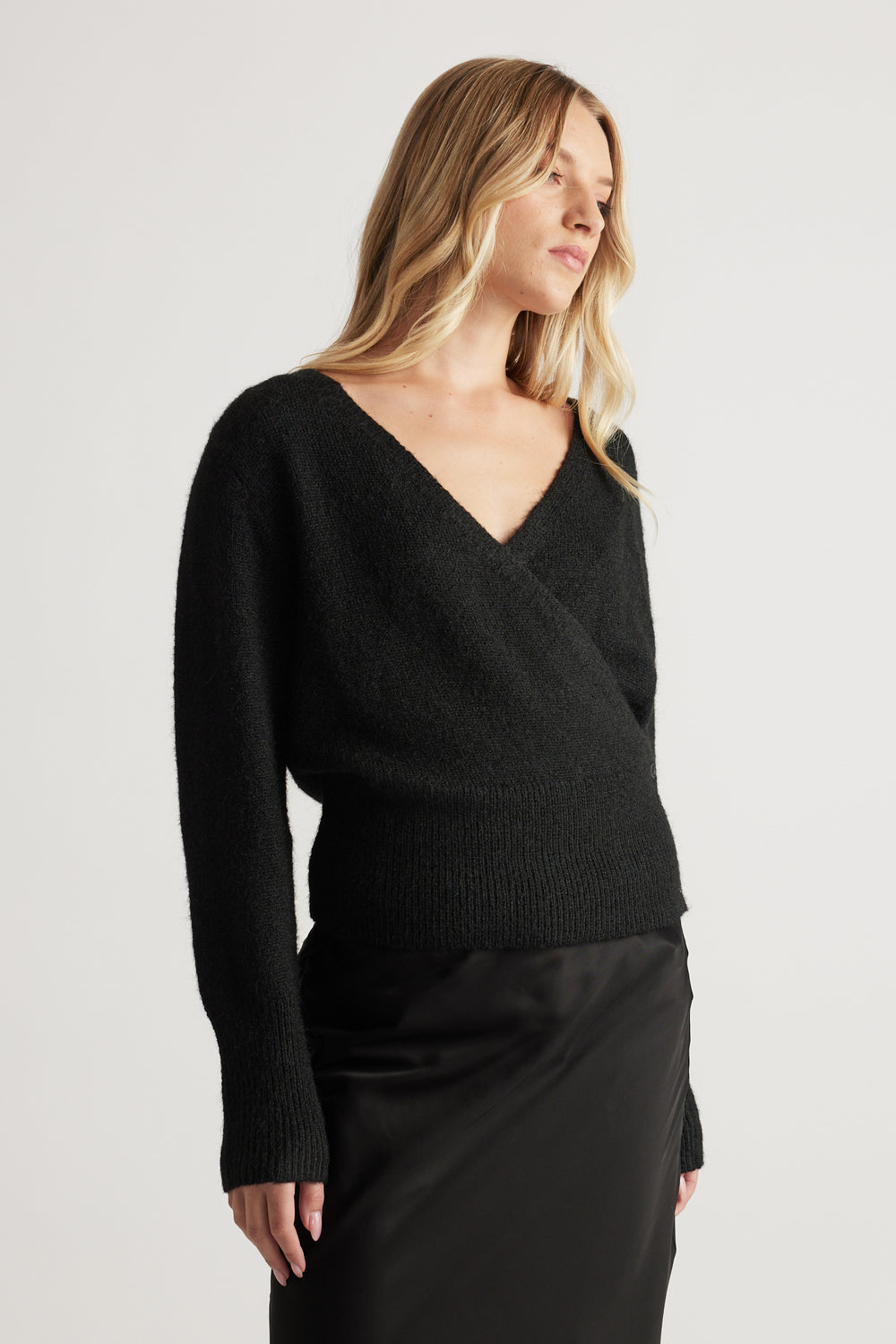 Woman wearing a soft black crossover knit cardigan with long sleeves paired with a sleek black skirt against a plain background.