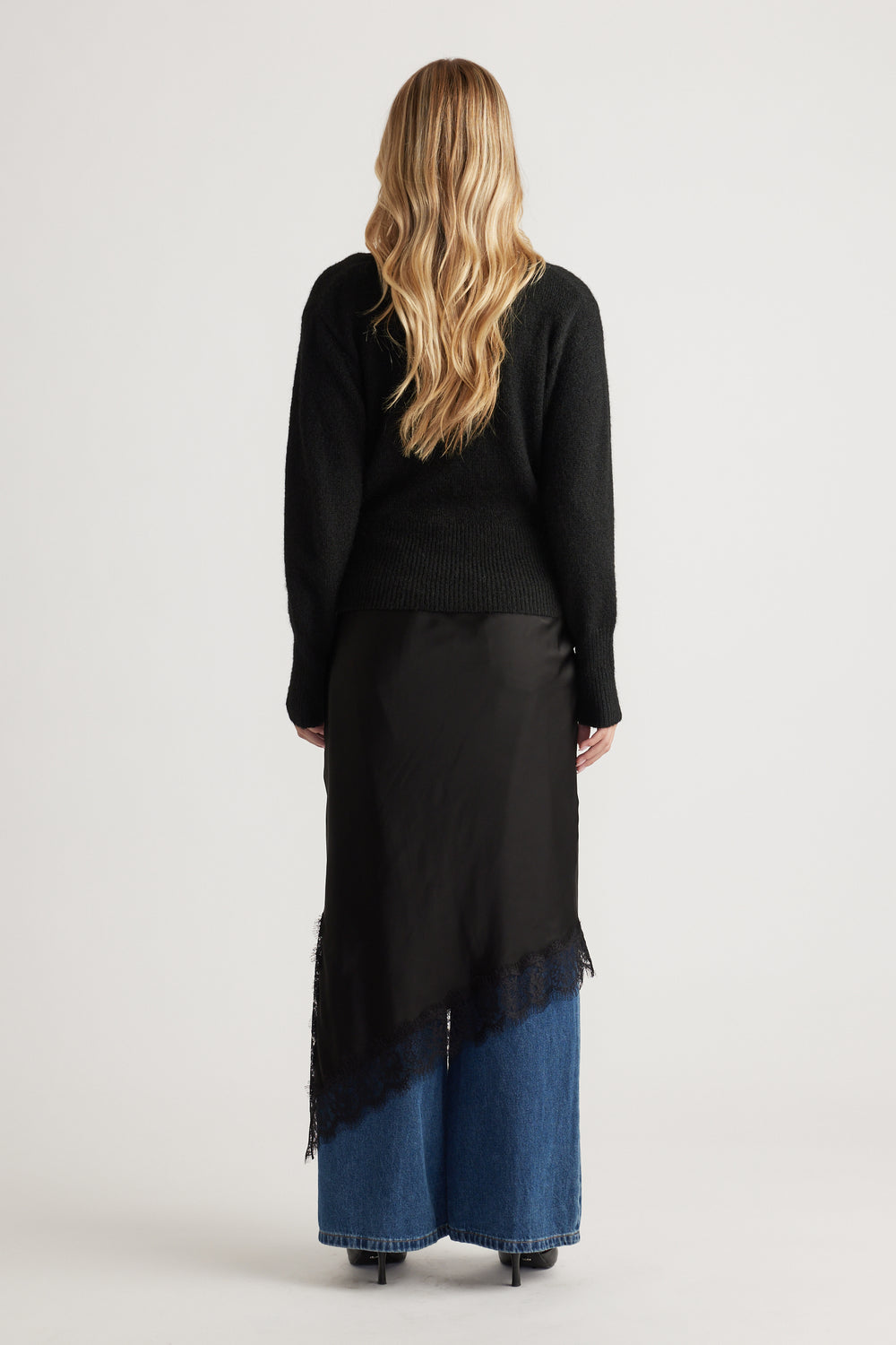 Back view of a black knit crossover cardigan layered over a silky asymmetrical lace-trimmed skirt paired with wide-leg blue denim jeans