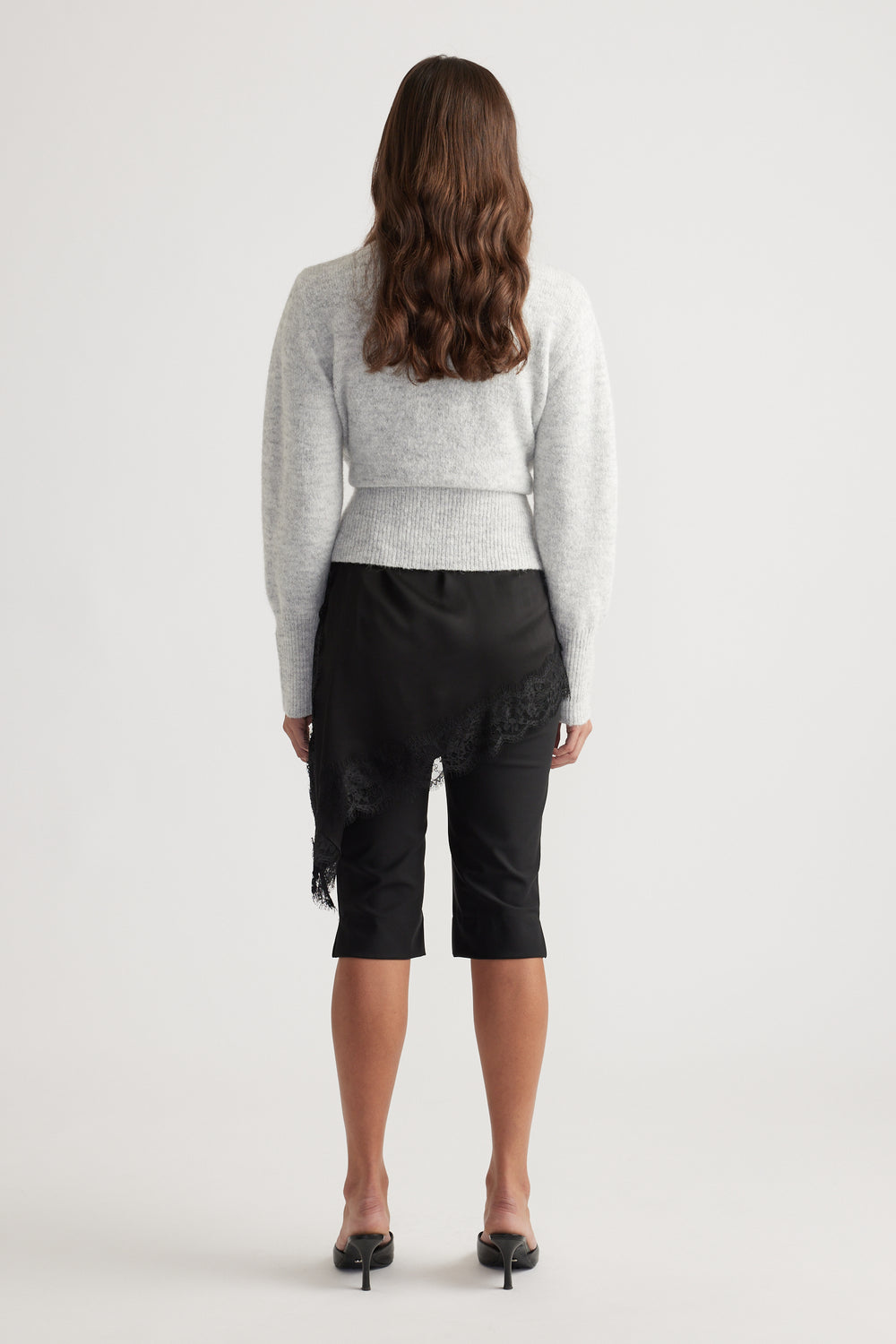 Back view of a soft grey knit crossover cardigan paired with a black asymmetrical lace-trimmed skirt and heels.