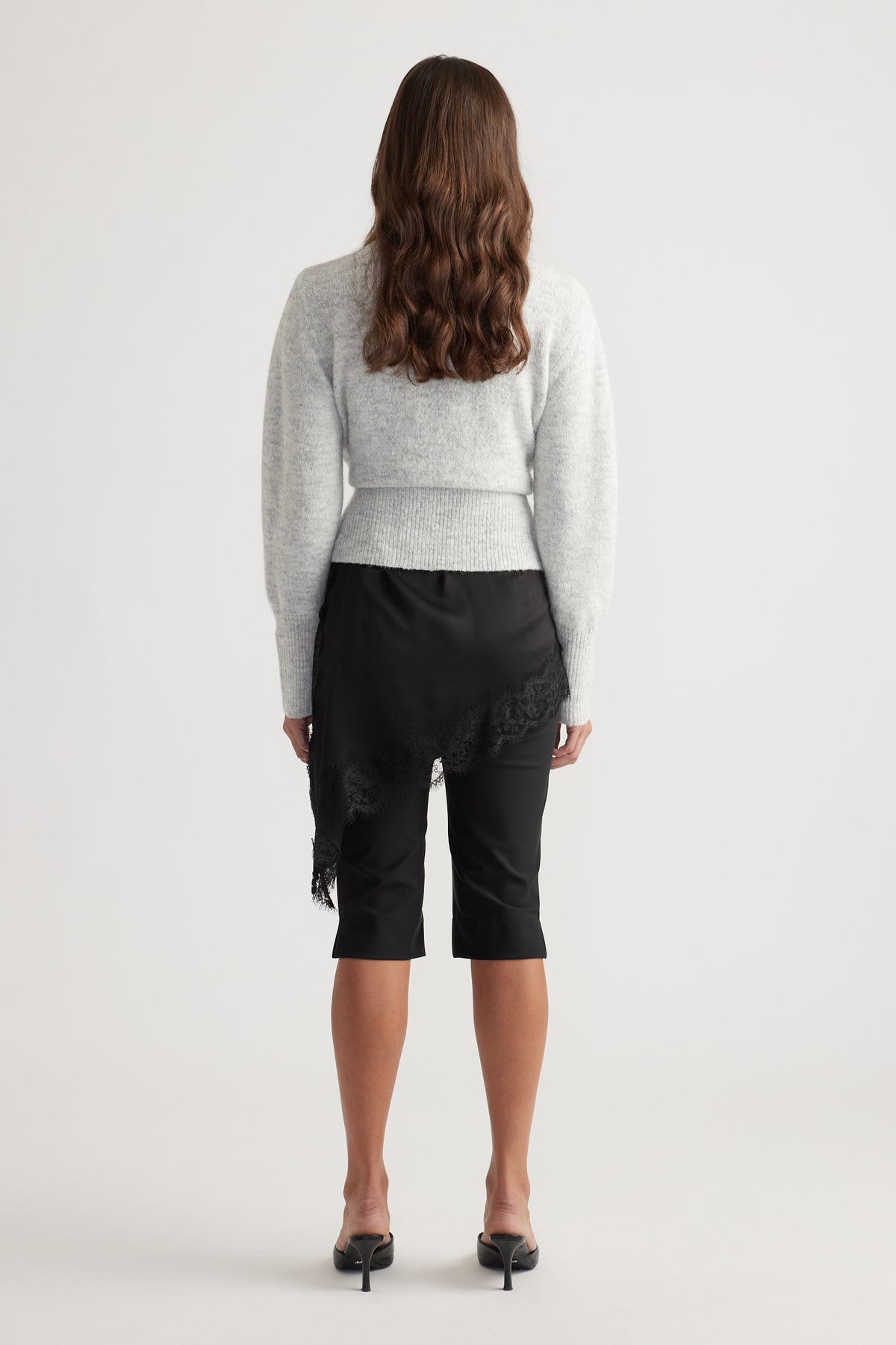 Back view of a soft grey knit crossover cardigan paired with a black asymmetrical lace-trimmed skirt and heels.