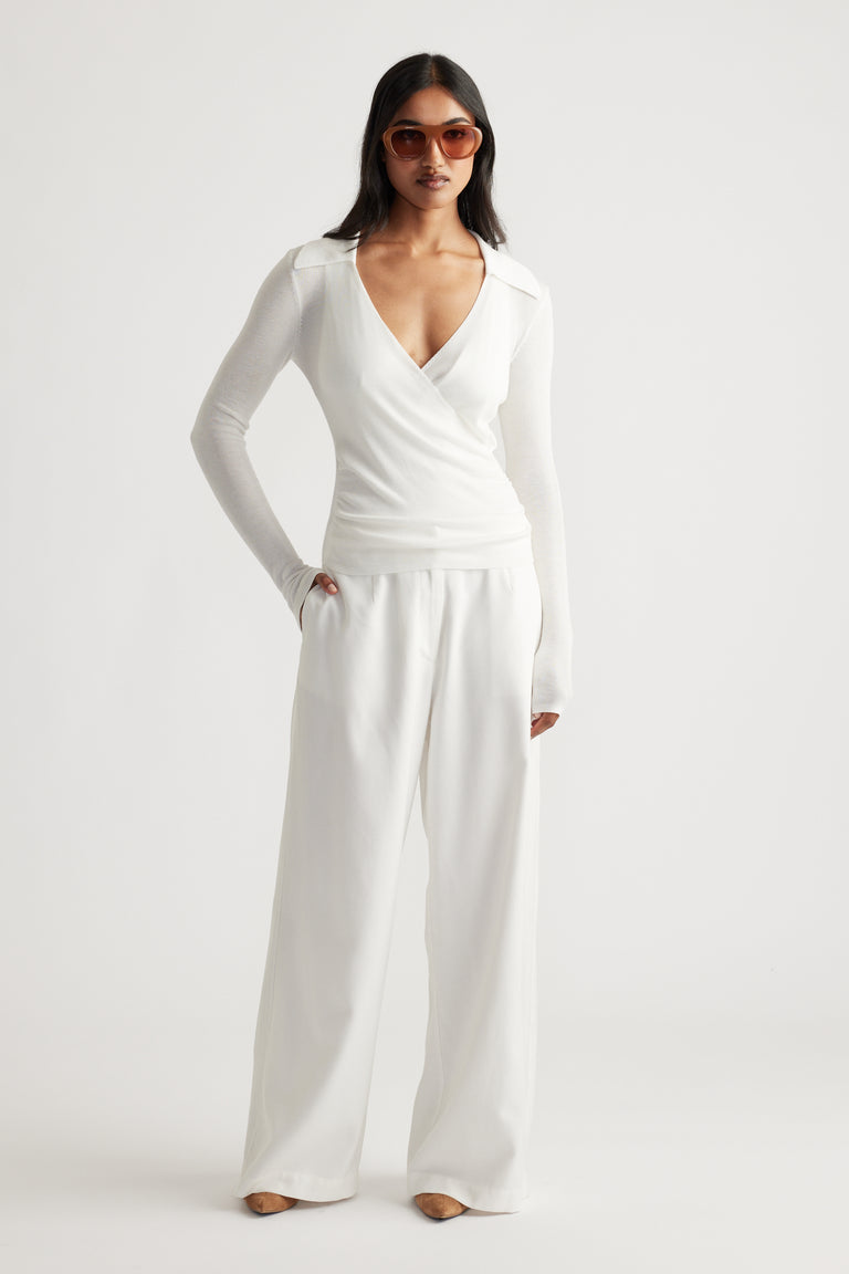 Model wearing an oyster-tone crossover knit top paired with wide-leg trousers and oversized sunglasses in a minimalist setting.