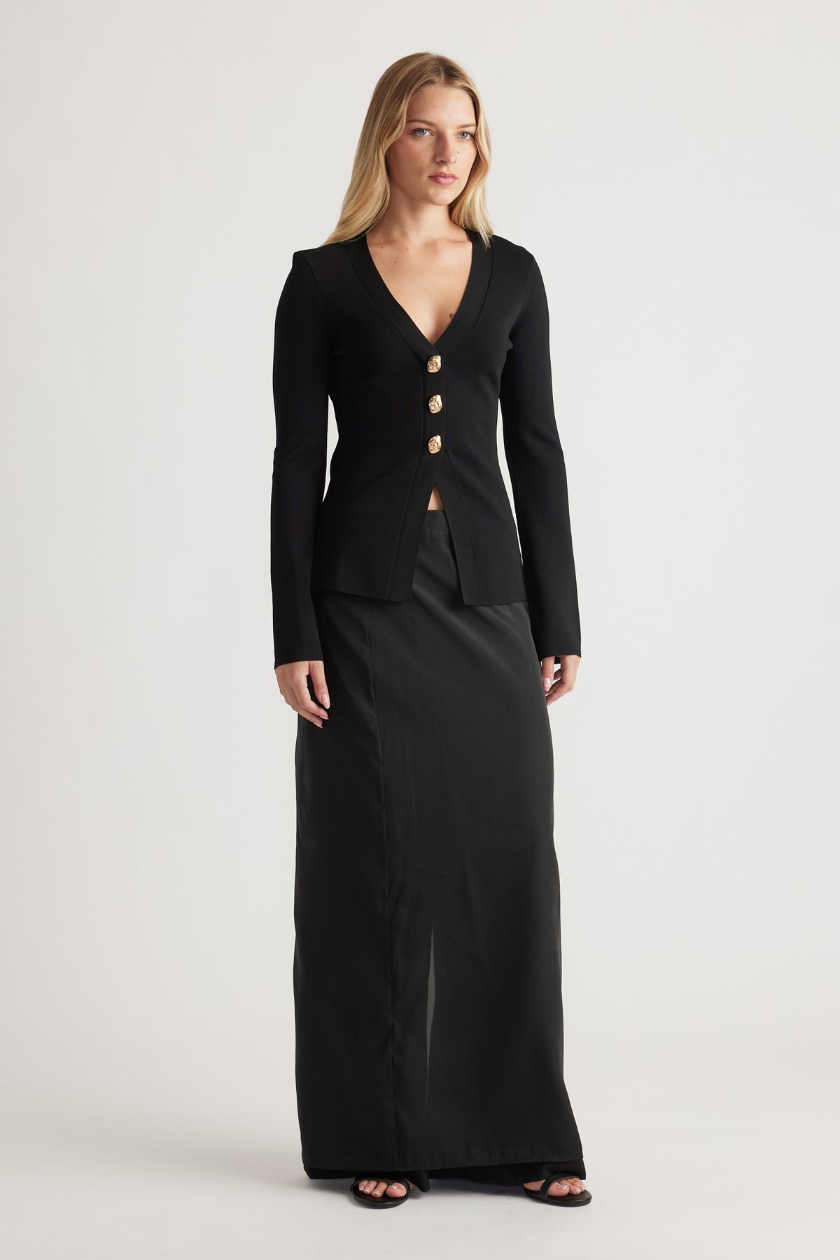 Woman wearing a fitted black knit cardigan with gold buttons and deep V-neck, paired with a long black skirt