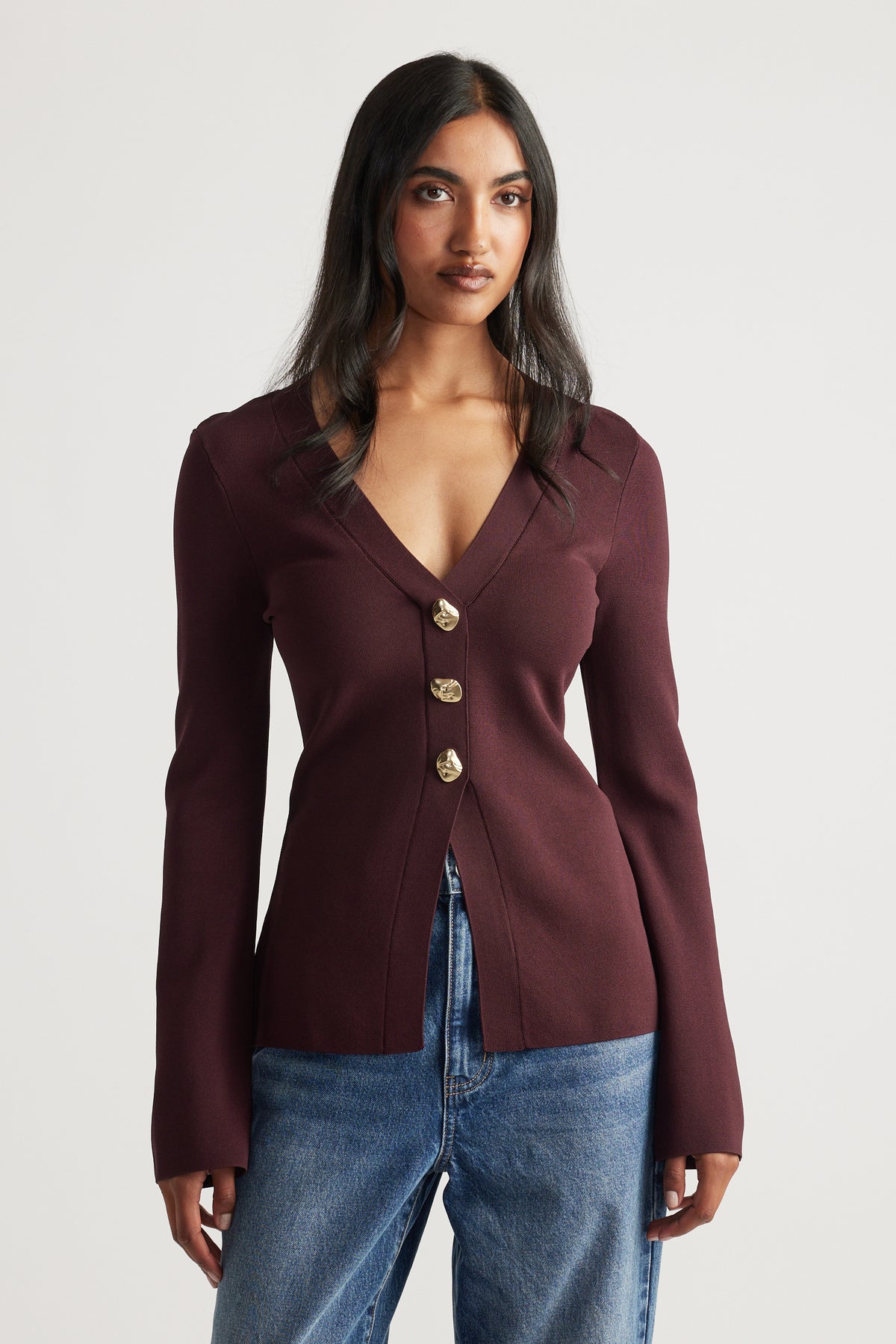 Front view of a woman wearing a fitted burgundy longline knit cardigan with gold buttons and relaxed blue jeans