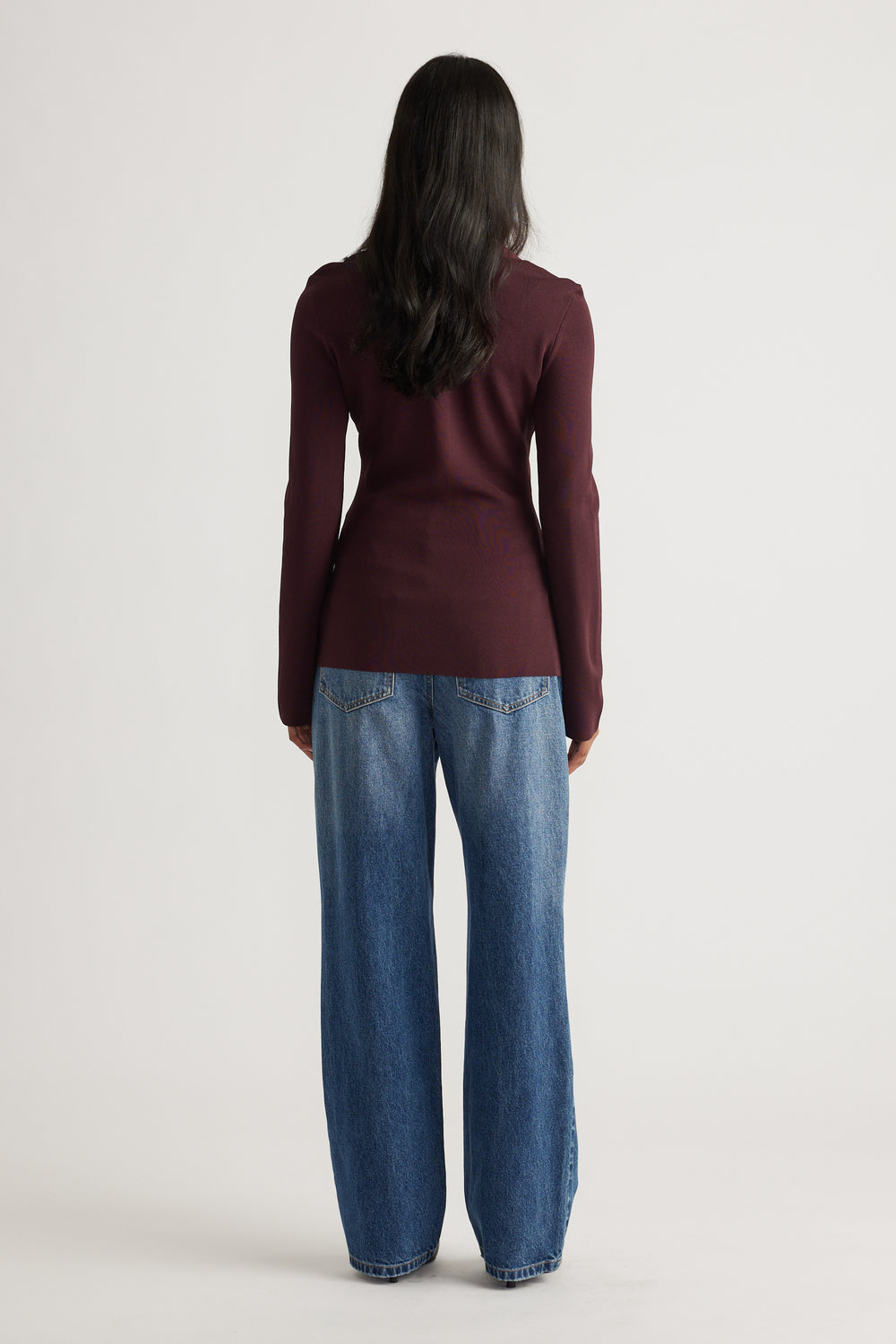 Woman seen from behind wearing a fitted long-sleeve burgundy knit top paired with relaxed wide-leg blue denim jeans