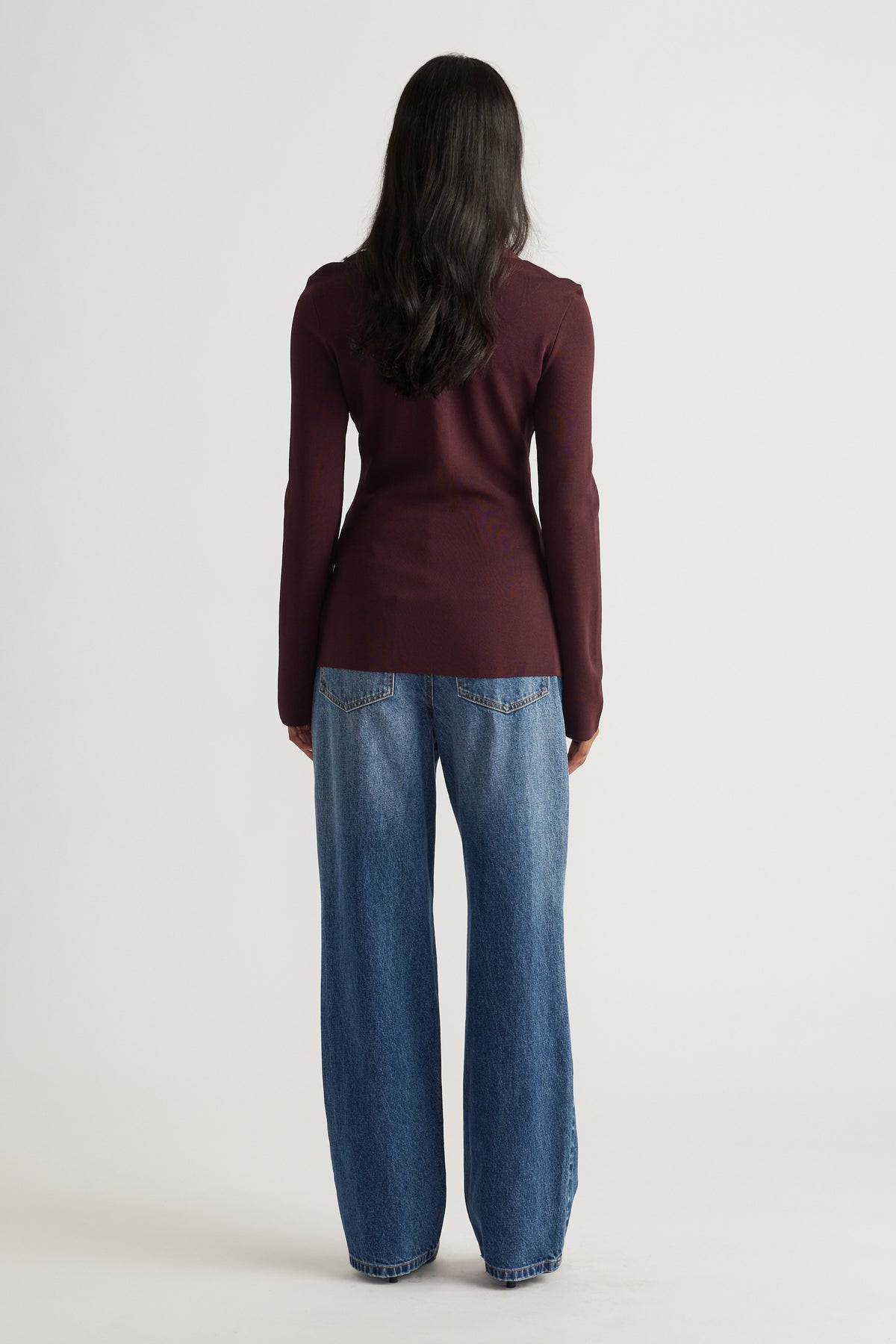 Woman seen from behind wearing a fitted long-sleeve burgundy knit top paired with relaxed wide-leg blue denim jeans