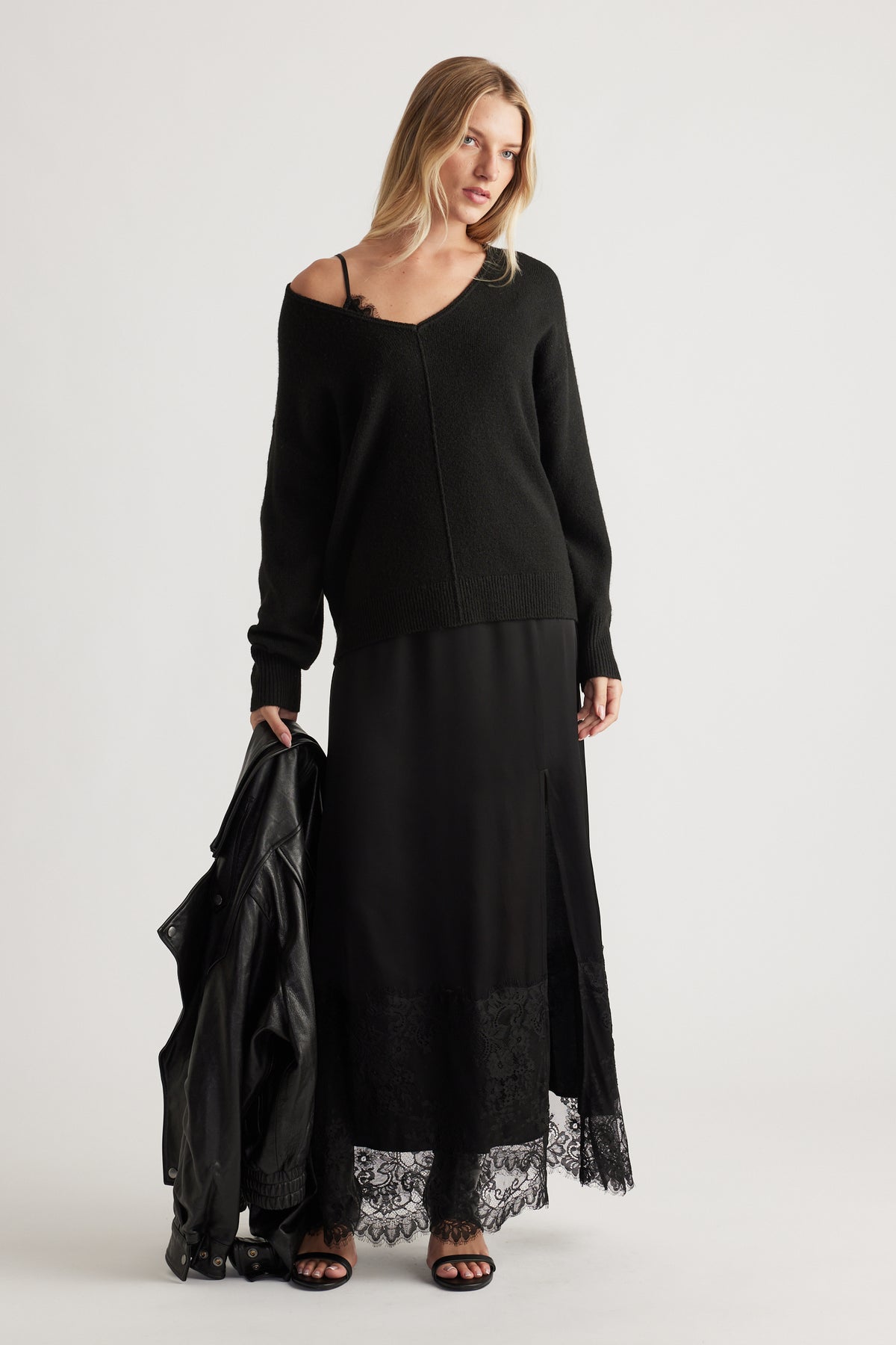 Woman wearing a relaxed black knit jumper with a central seam, paired with a long black skirt featuring lace trim and a side slit.
