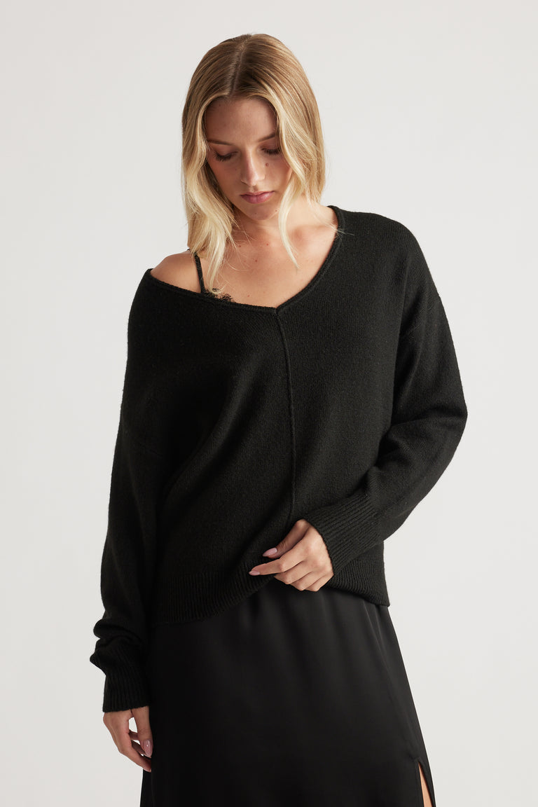Woman wearing a relaxed black knit jumper with a central seam detail, paired with a black skirt featuring side slits