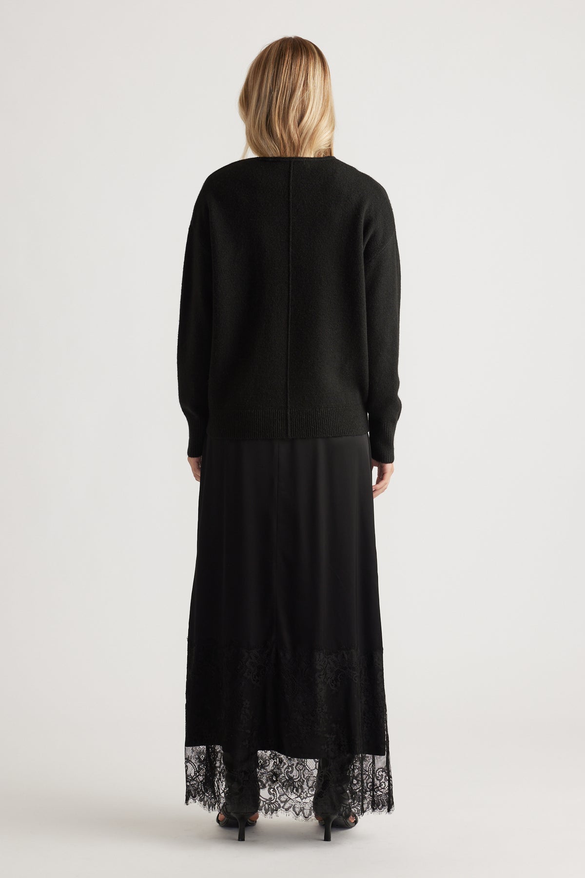 Back view of woman wearing a black knit jumper with seam detail paired with a long black lace-trimmed skirt and heels