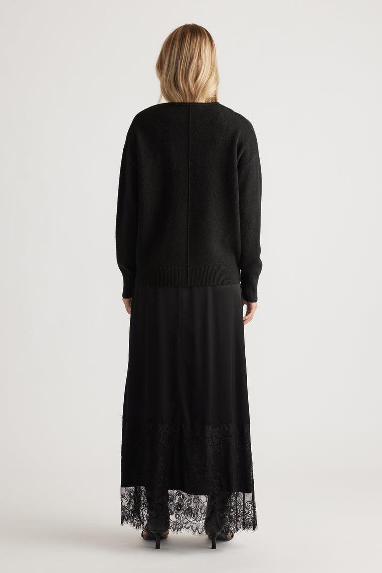 Back view of woman wearing a black knit jumper with seam detail paired with a long black lace-trimmed skirt and heels
