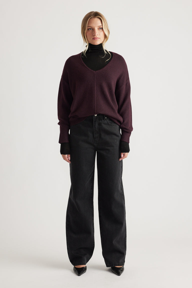Woman wearing a relaxed-fit burgundy V-neck knit jumper with central seam over a black turtleneck and wide-leg black jeans