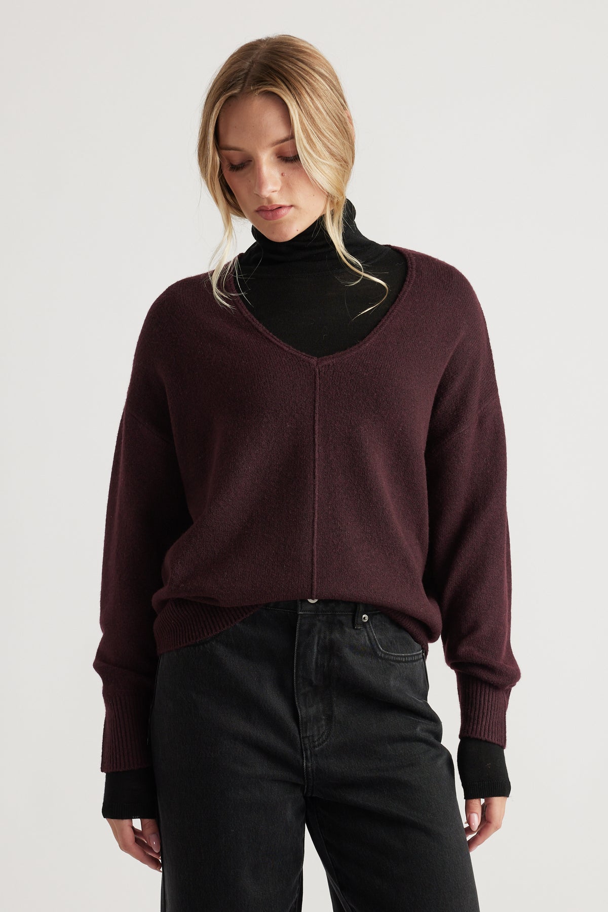 Woman wearing a deep burgundy knit jumper with seam detail over a black turtleneck and high-waisted black jeans
