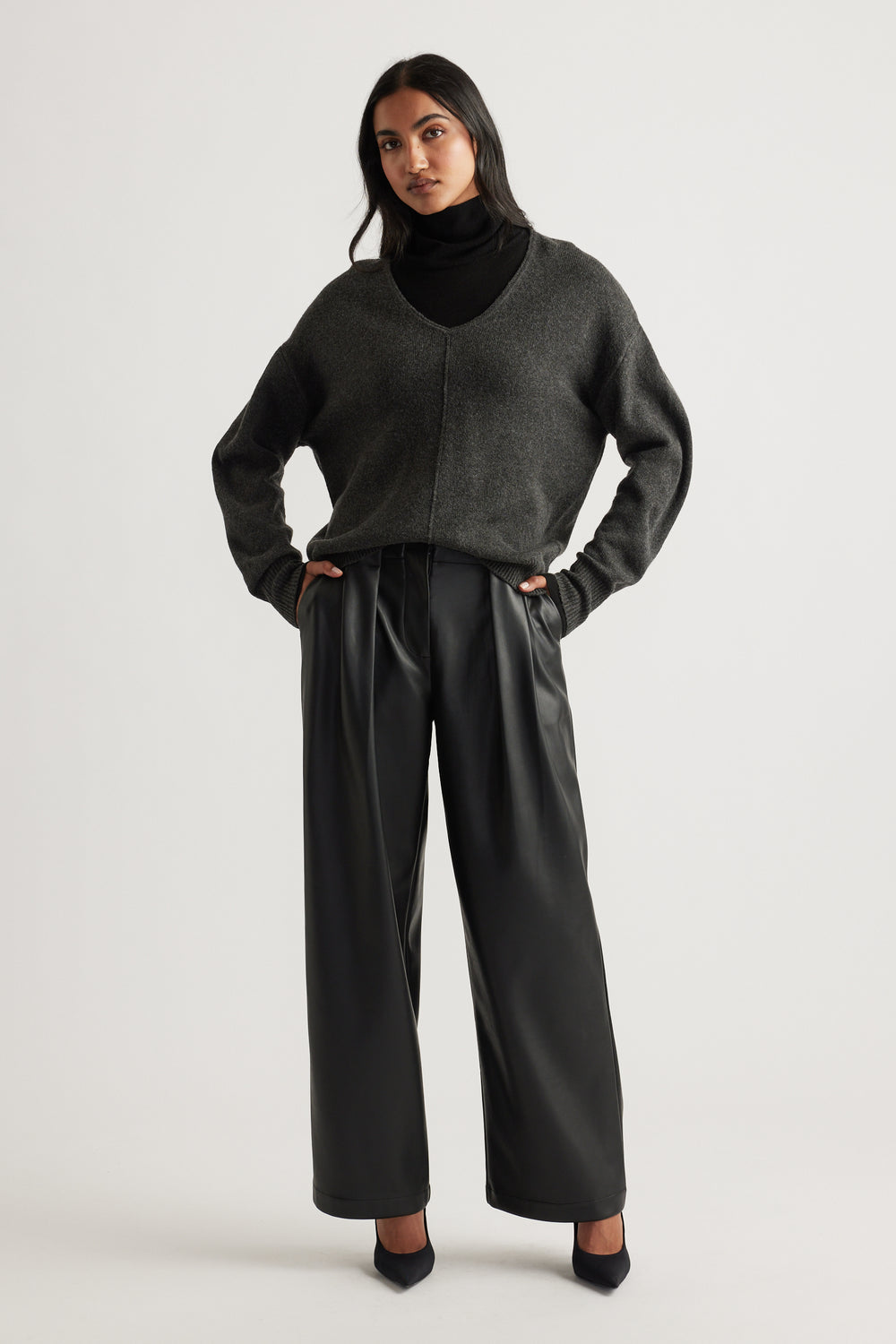Woman wearing a charcoal knit jumper with a deep V-neck and seam detail, paired with black wide-leg trousers and heels