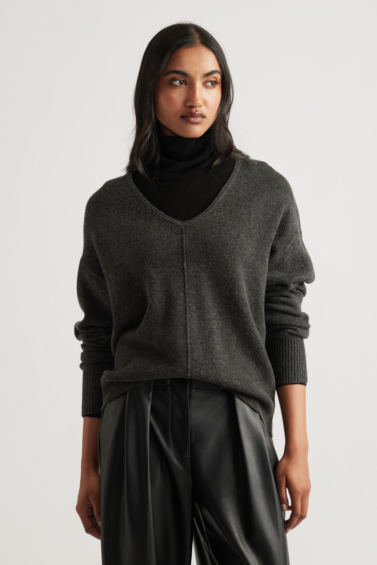 Woman wearing a charcoal knit jumper with a V-neck seam detail layered over a black turtleneck and black pants