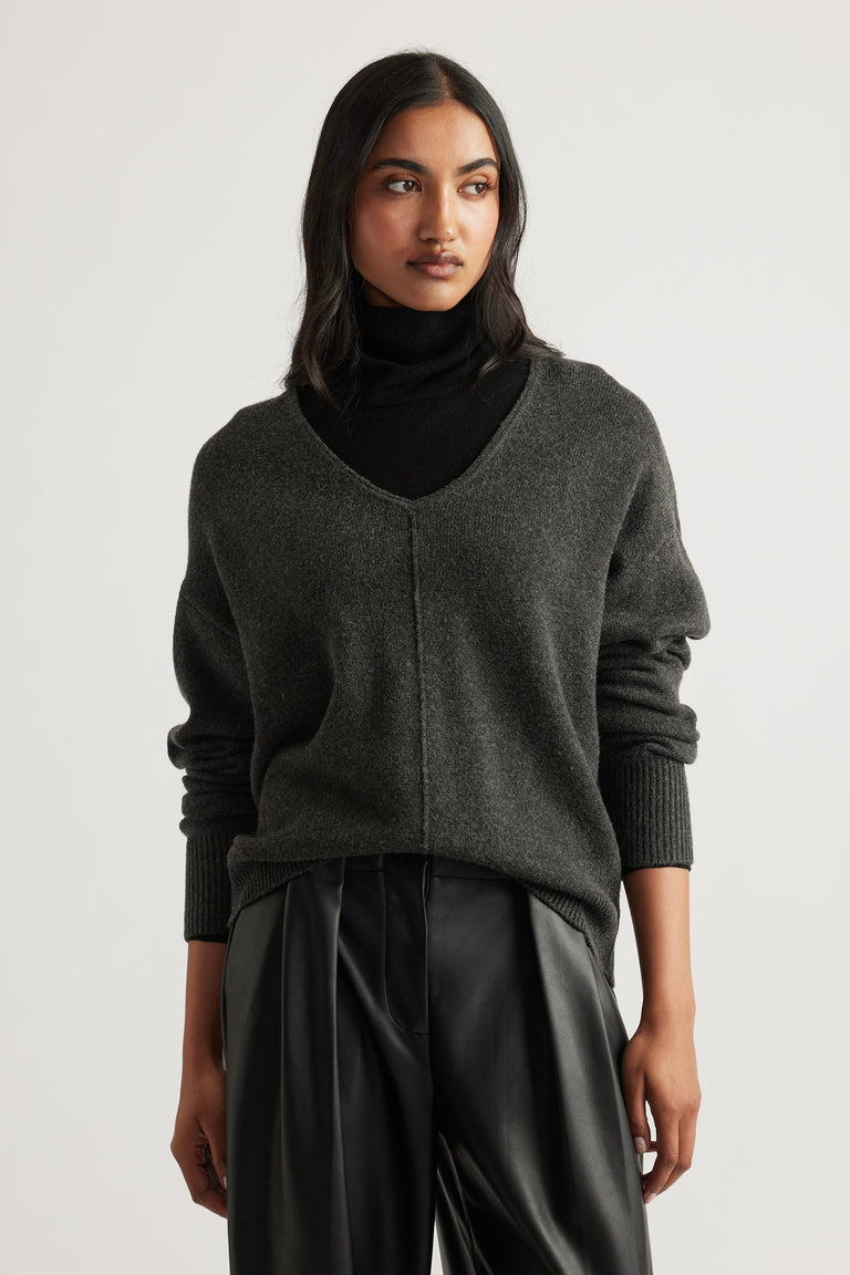 Woman wearing a charcoal knit jumper with a V-neck seam detail layered over a black turtleneck and black pants