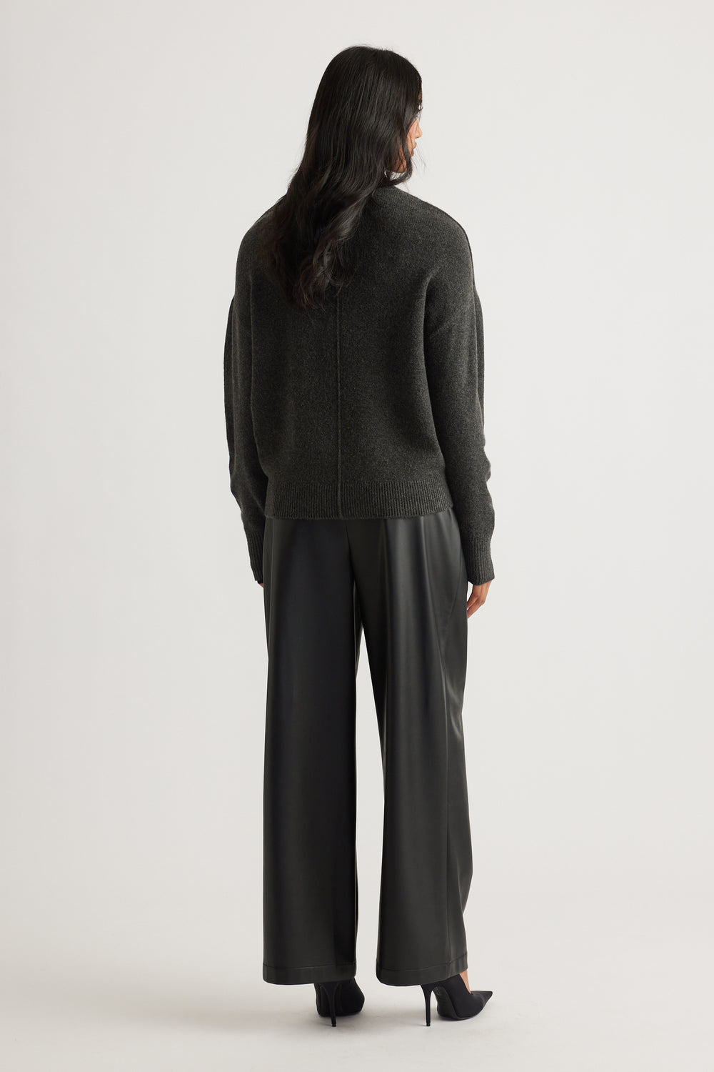 Woman wearing a charcoal knit jumper with subtle seam detail, paired with black wide-leg leather-look pants and heels