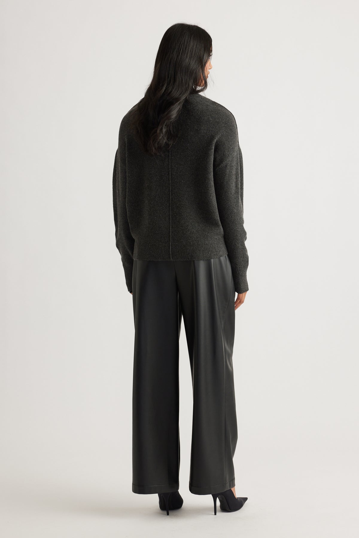 Woman wearing a charcoal knit jumper with subtle seam detail, paired with black wide-leg leather-look pants and heels