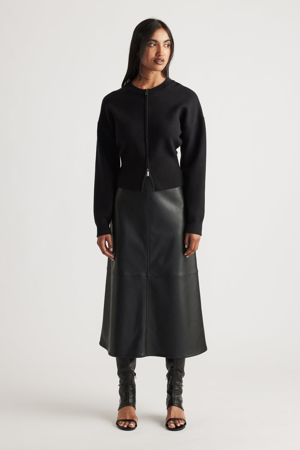 Woman wearing black knit bomber jacket with zip front, paired with a midi faux leather skirt and open-toe boots