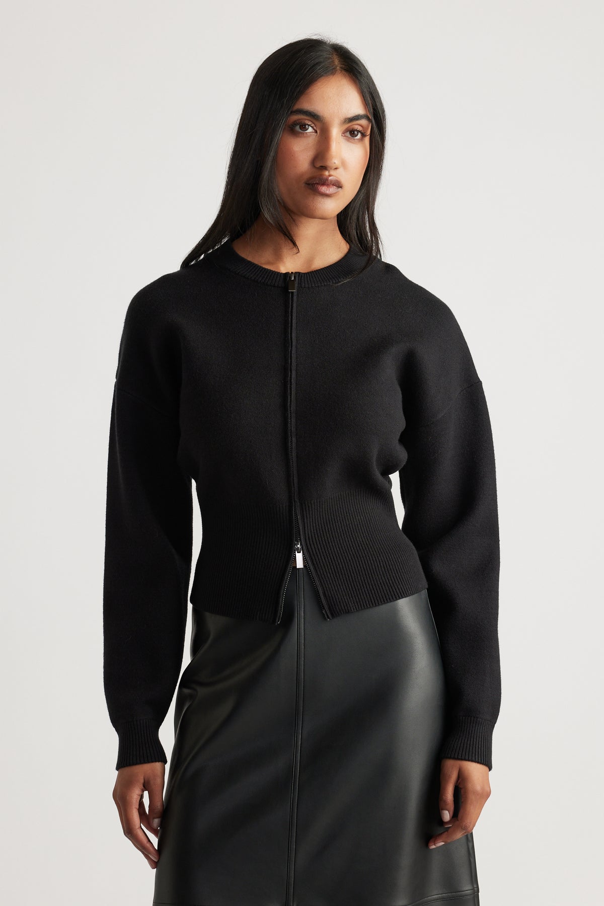 Woman wearing a black zip-up knit bomber with ribbed waist and cuffs, paired with a black leather-look skirt