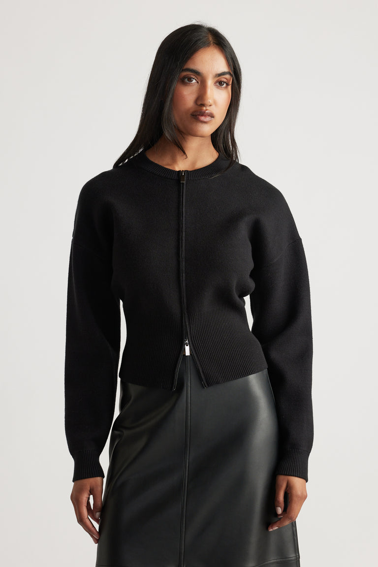 Woman wearing a black zip-up knit bomber with ribbed waist and cuffs, paired with a black leather-look skirt
