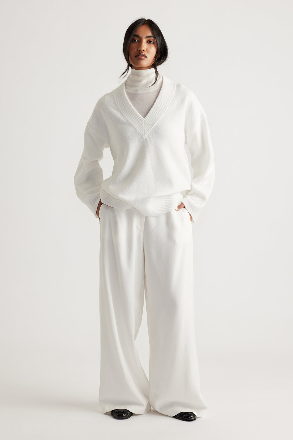 Model dressed in a soft white V-neck knit jumper layered over a high-neck top, paired with wide-leg trousers.