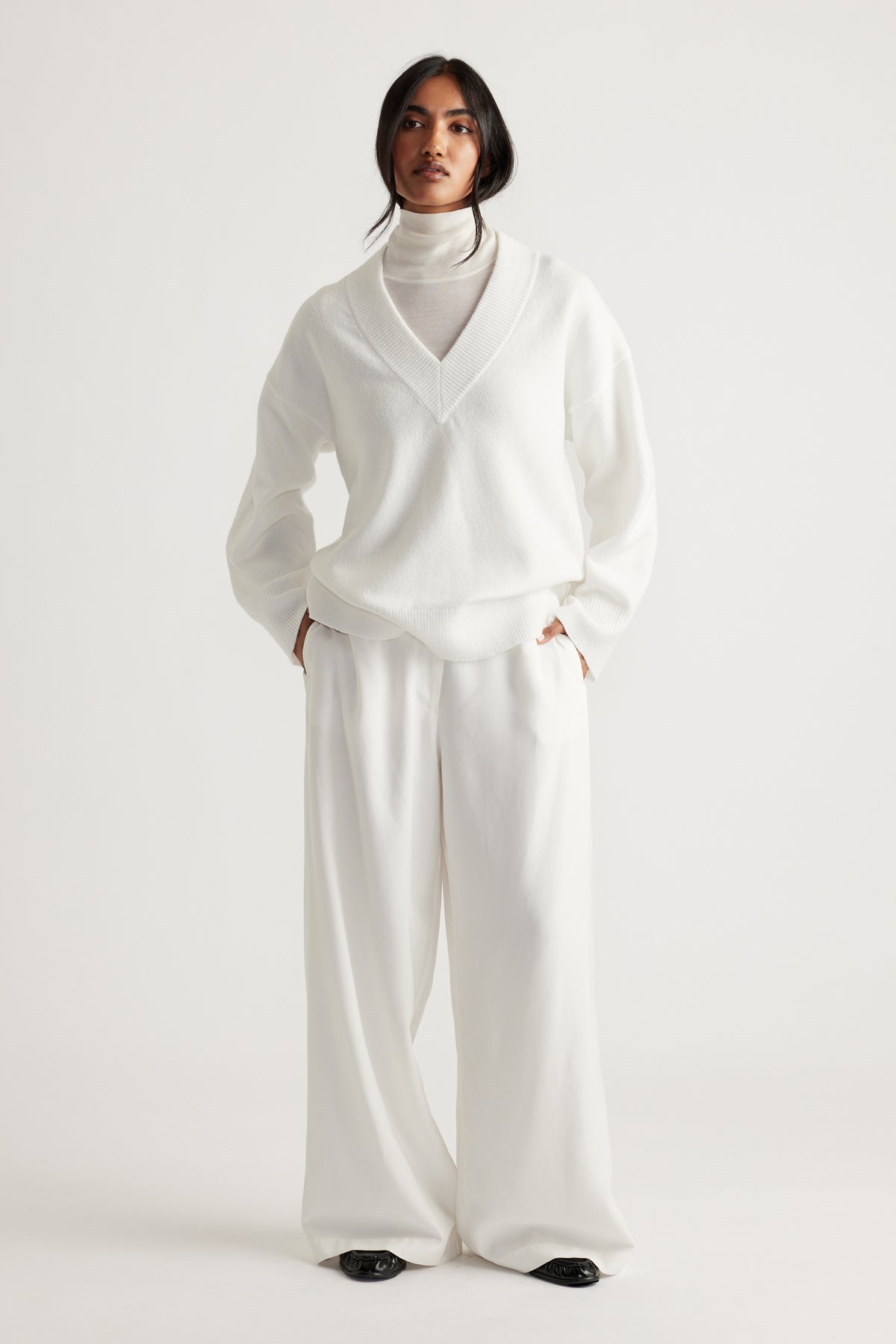 Model dressed in a soft white V-neck knit jumper layered over a high-neck top, paired with wide-leg trousers.