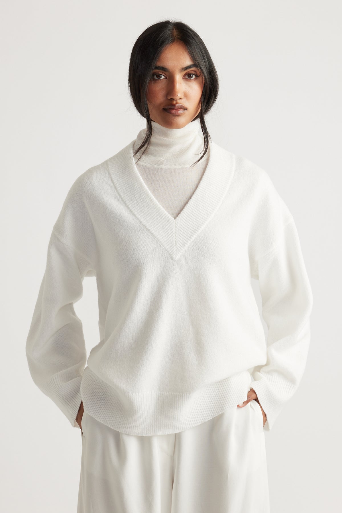 Woman wearing a soft white V-neck knit jumper layered over a light turtleneck, styled with relaxed white pants for a clean look