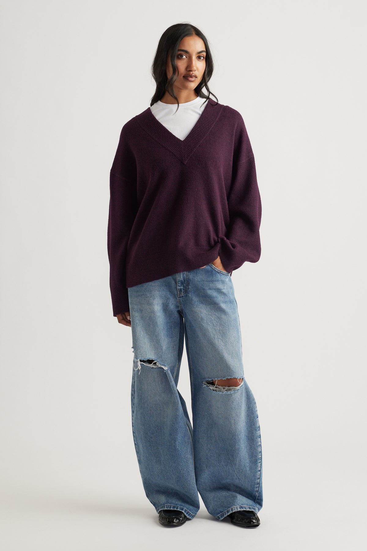 Woman wearing a deep plum V-neck knit jumper layered over a white tee, paired with relaxed wide-leg ripped jeans