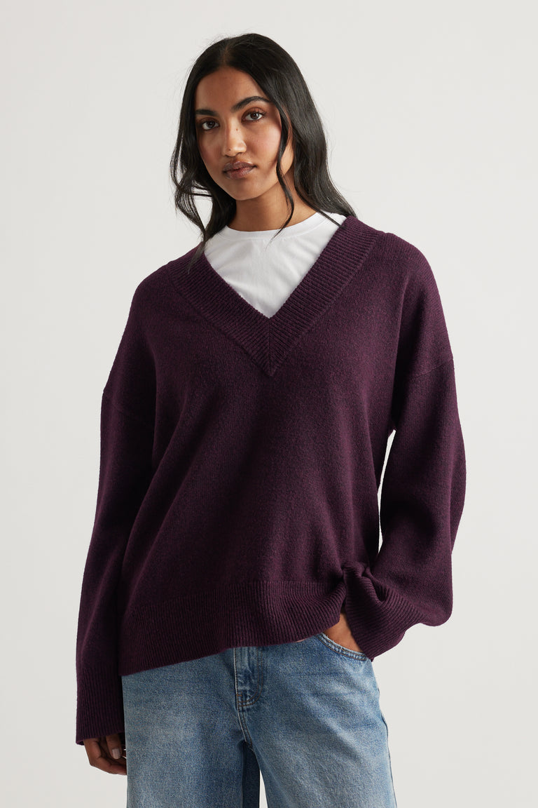 Woman wearing a deep plum oversized V-neck knit jumper layered over a white tee, paired with relaxed blue jeans