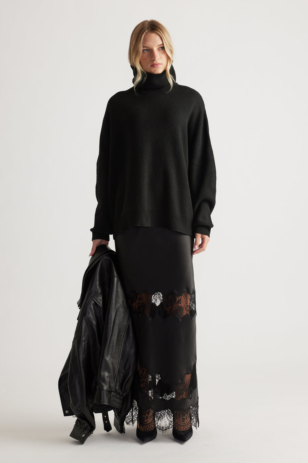 Woman wearing a black roll neck knit jumper with loose sleeves paired with a black lace-trimmed maxi skirt and leather jacket