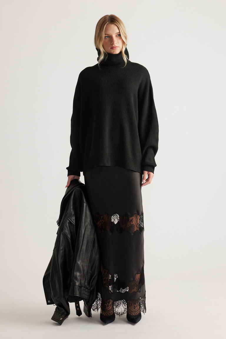Woman wearing a black roll neck knit jumper with loose sleeves paired with a black lace-trimmed maxi skirt and leather jacket