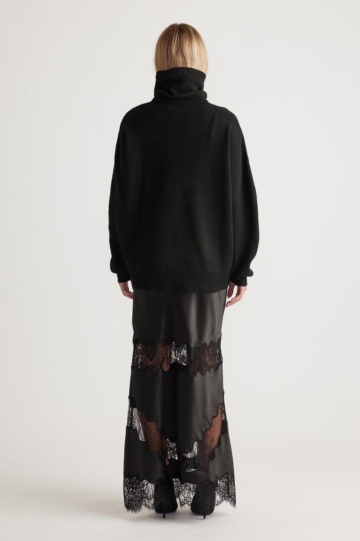 Woman seen from behind wearing a black roll neck knit jumper paired with a black satin maxi skirt with lace panels