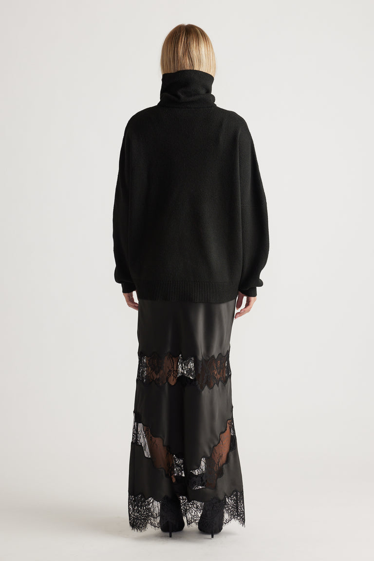 Woman seen from behind wearing a black roll neck knit jumper paired with a black satin maxi skirt with lace panels