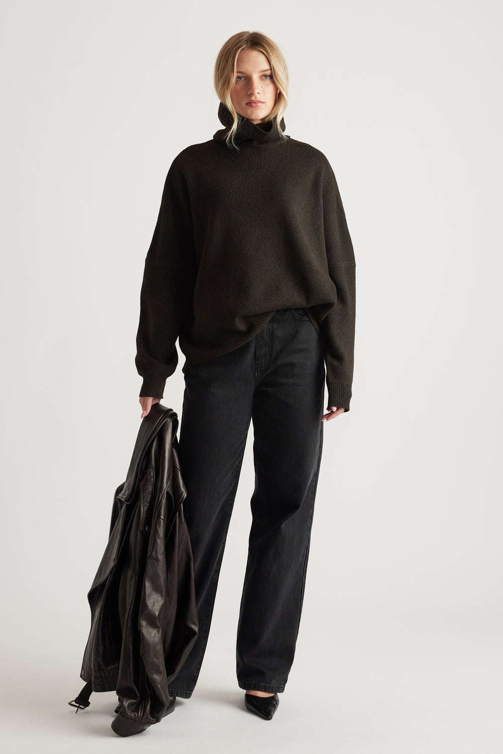 Woman wearing a dark roll neck knit jumper paired with relaxed black jeans and holding a leather jacket for a casual look