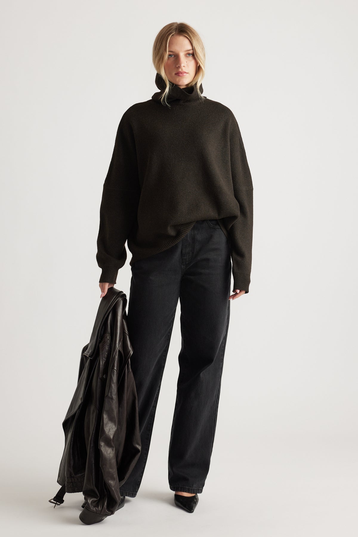 Woman wearing a dark roll neck knit jumper paired with relaxed black jeans and holding a leather jacket for a casual look