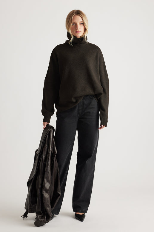 Woman wearing a dark roll neck knit jumper paired with relaxed black jeans and holding a leather jacket for a casual look