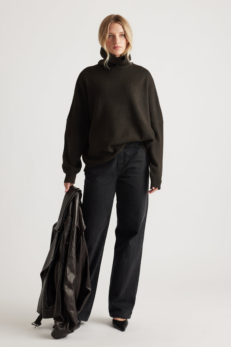 Woman wearing a dark roll neck knit jumper paired with relaxed black jeans and holding a leather jacket for a casual look