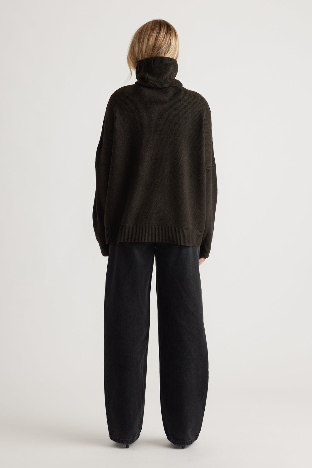 Woman wearing a loose dark brown roll neck jumper paired with relaxed black wide-leg pants in a minimalist setting