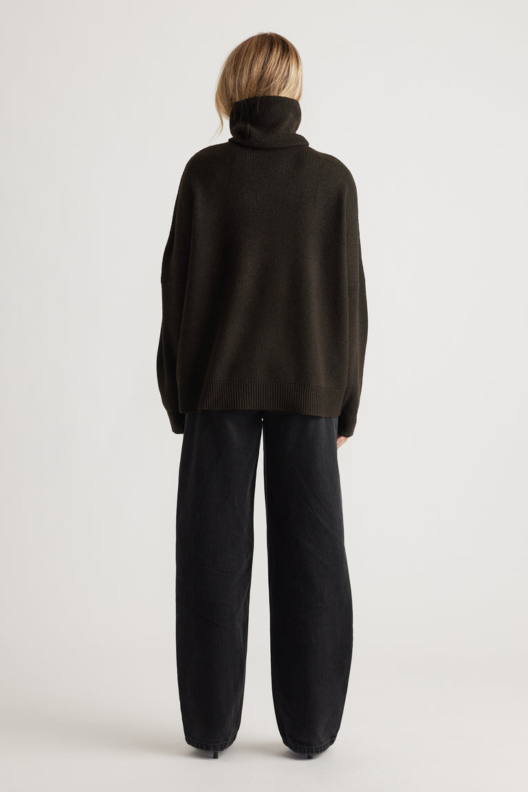 Woman wearing a loose dark brown roll neck jumper paired with relaxed black wide-leg pants in a minimalist setting